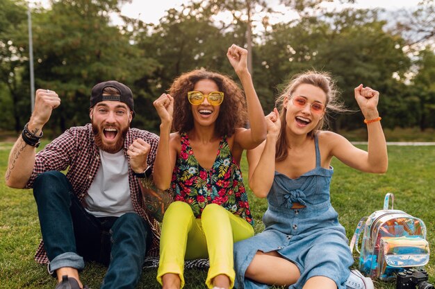 Felice giovane compagnia di amici sorridenti seduti nel parco su erba, uomini e donne che si divertono insieme