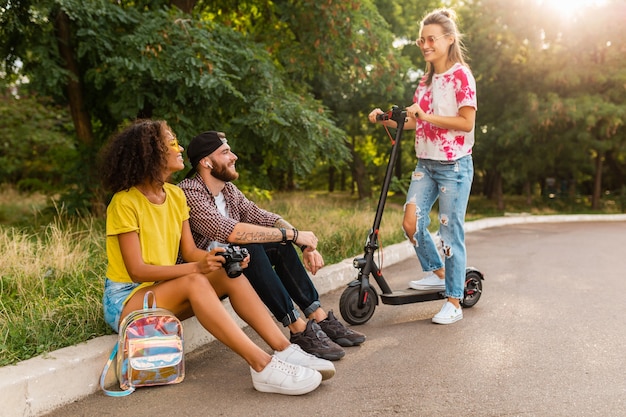 Felice giovane compagnia di amici sorridenti seduti nel parco sull'erba con scooter elettrico, uomini e donne che si divertono insieme