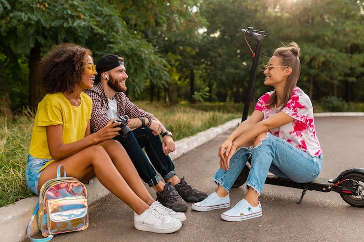 Foto gratuito felice giovane compagnia di amici sorridenti seduti nel parco sull'erba con scooter elettrico, uomini e donne che si divertono insieme