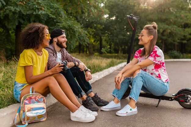 Felice giovane compagnia di amici sorridenti seduti nel parco sull'erba con scooter elettrico, uomini e donne che si divertono insieme