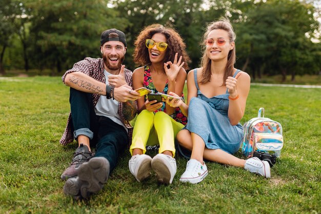 Felice giovane compagnia di amici sorridenti seduti nel parco utilizzando smartphone, uomini e donne che hanno divertimento insieme