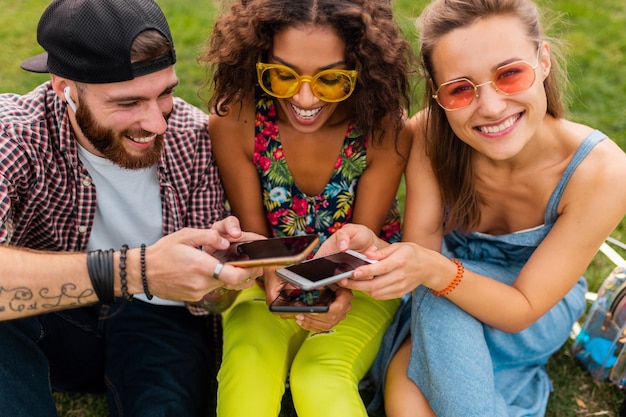 Felice giovane compagnia di amici sorridenti seduti nel parco utilizzando smartphone, uomini e donne che hanno divertimento insieme