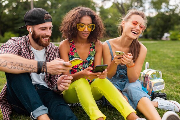 Felice giovane compagnia di amici sorridenti seduti nel parco utilizzando smartphone, uomini e donne che hanno divertimento insieme