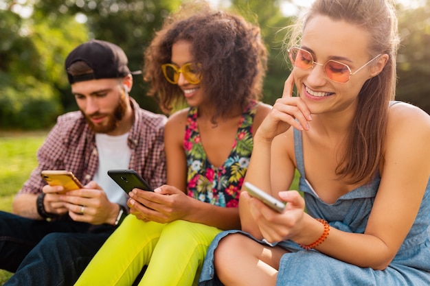 Felice giovane compagnia di amici sorridenti seduti nel parco utilizzando smartphone, uomini e donne che hanno divertimento insieme