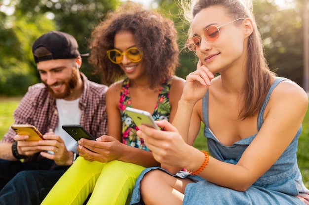 Felice giovane compagnia di amici sorridenti seduti nel parco utilizzando smartphone, uomini e donne che hanno divertimento insieme