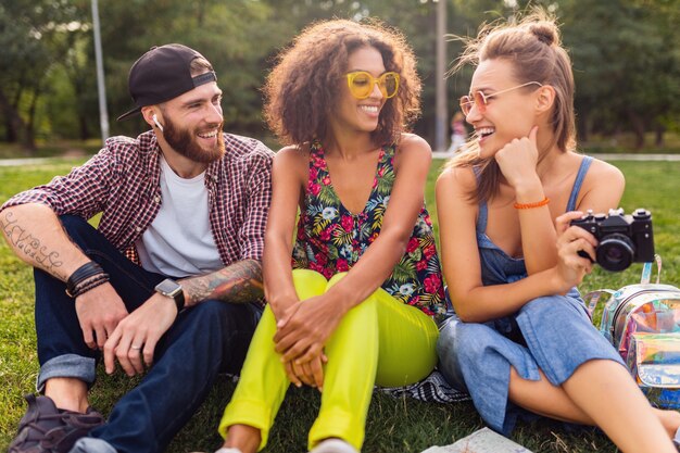 Felice giovane compagnia di parlare amici sorridenti seduti al parco, uomo e donna che si divertono insieme, colorato stile moda hipster estate, viaggiando con la macchina fotografica