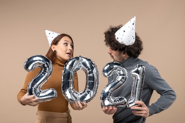 Felice giovane coppia in cerca di ogni altro indossare il cappello del nuovo anno pone per la fotocamera Ragazza che mostra ee ragazzo con e su grigio