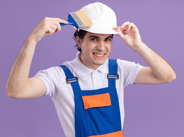 Felice giovane costruttore in uniforme da costruzione e casco di sicurezza che tiene il pennello guardando davanti sorridente in piedi sopra la parete viola