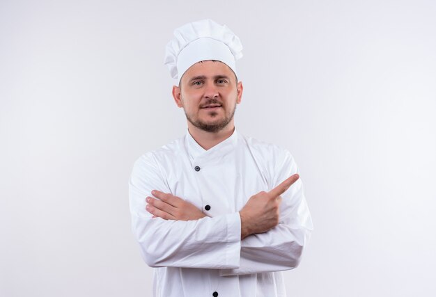 Felice giovane cuoco bello in uniforme da chef in piedi con la postura chiusa che punta a destra isolato su uno spazio bianco