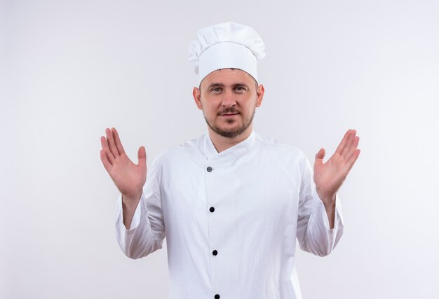 Felice giovane cuoco bello in uniforme del cuoco unico che mostra le mani vuote isolate su uno spazio bianco