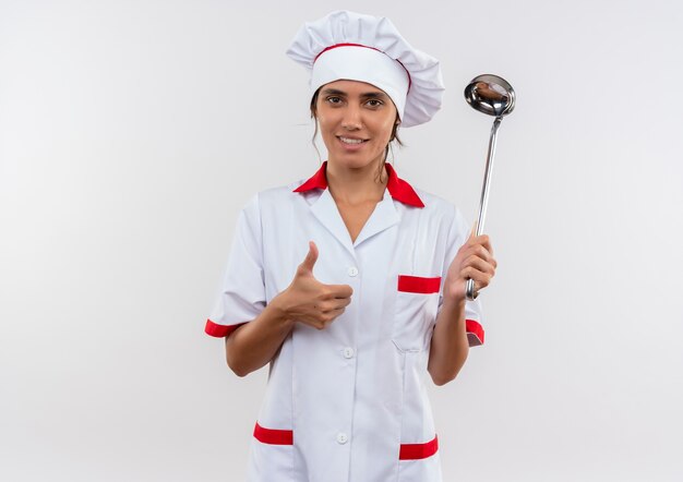 Felice giovane cuoco femminile che indossa uniforme chef tenendo mestolo il suo pollice in alto sulla parete bianca isolata con lo spazio della copia