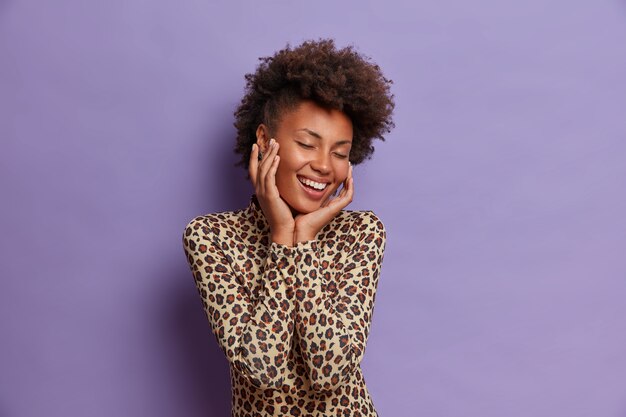 Felice giovane donna afroamericana spensierata con bellezza naturale, capelli ricci, ampio sorriso piacevole, tocca il viso, gode di una pelle morbida, chiude gli occhi con soddisfazione