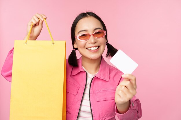 Felice giovane donna asiatica che mostra la carta di credito per lo shopping tenendo la borsa che acquista in vendita andando al negozio in piedi su sfondo rosa