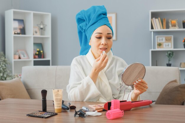 Felice giovane donna asiatica con un asciugamano in testa seduto al tavolo da toeletta a casa interno guardando lo specchio applicando il rossetto facendo routine di trucco mattutino