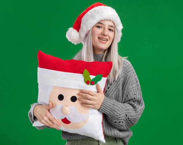 Felice giovane donna bionda in maglione invernale e cappello santa tenendo il cuscino di Natale guardando la fotocamera con il sorriso sul viso in piedi su sfondo verde