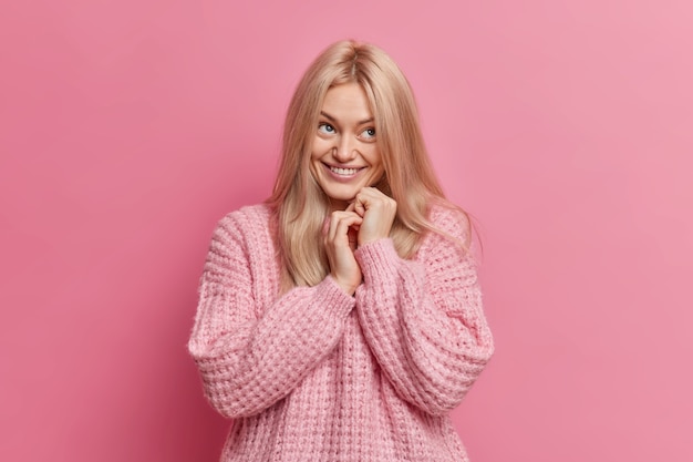 felice giovane donna bionda tiene le mani unite e ha un'espressione del viso positivo sognante vestito con un maglione lavorato a maglia sciolto