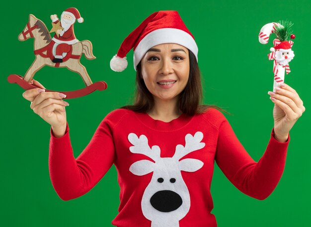felice giovane donna che indossa il cappello di Babbo Natale e maglione rosso in possesso di giocattoli di Natale guardando la fotocamera con il sorriso sul viso in piedi su sfondo verde