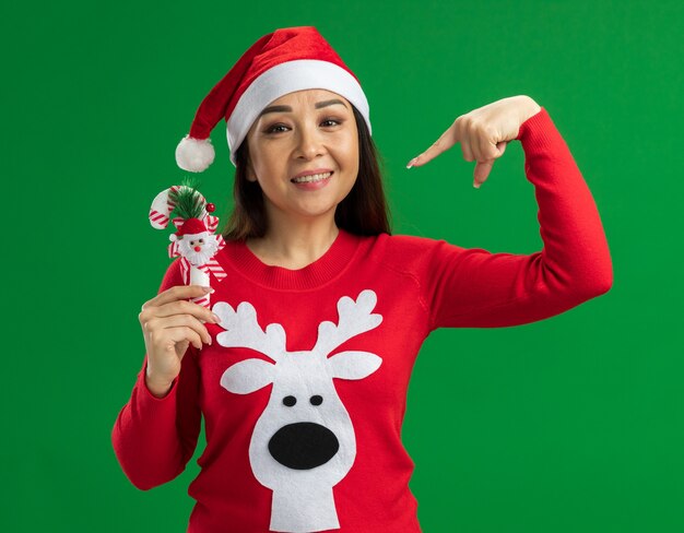 felice giovane donna che indossa il cappello di Babbo Natale e maglione rosso tenendo il bastoncino di zucchero di Natale che punta con il dito indice a sorridere in piedi su sfondo verde