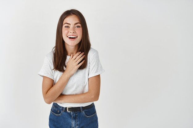 Felice giovane donna che ringrazia, sorride e indica se stessa, viene lodata e si congratula, in piedi sul muro bianco