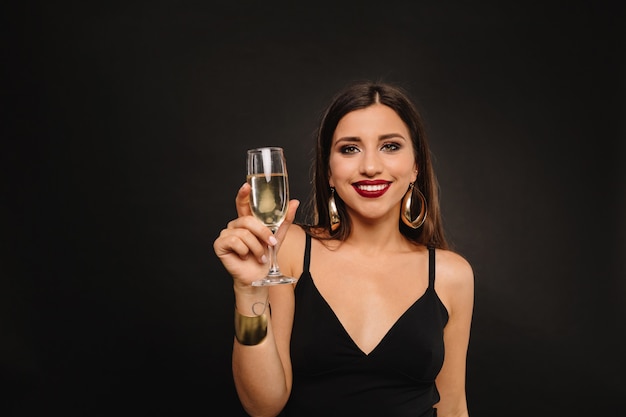 Felice giovane donna con gioielli d'oro in abito nero bevendo champagne