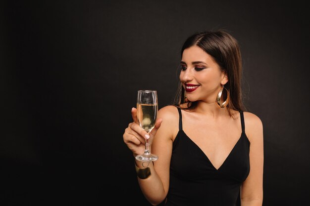 Felice giovane donna con gioielli d'oro in abito nero bevendo champagne
