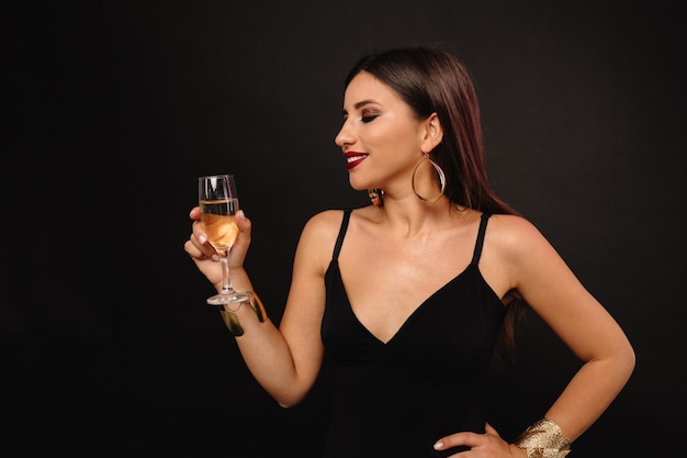 Felice giovane donna con gioielli d'oro in abito nero bevendo champagne