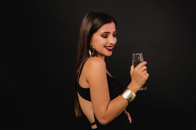 Felice giovane donna con gioielli d'oro in abito nero bevendo champagne