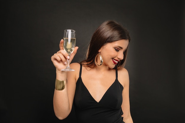 Felice giovane donna con gioielli d'oro in abito nero bevendo champagne