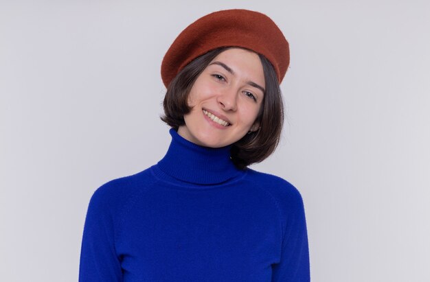 Felice giovane donna con i capelli corti in dolcevita blu che indossa berretto guardando davanti sorridendo allegramente in piedi sopra il muro bianco