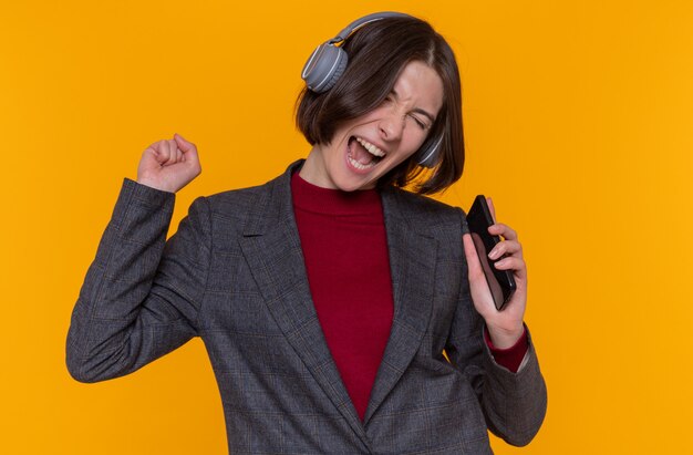 Felice giovane donna con i capelli corti indossa giacca grigia con le cuffie godendo la sua musica preferita cantando tenendo lo smartphone utilizzando come microfono in piedi sopra la parete arancione