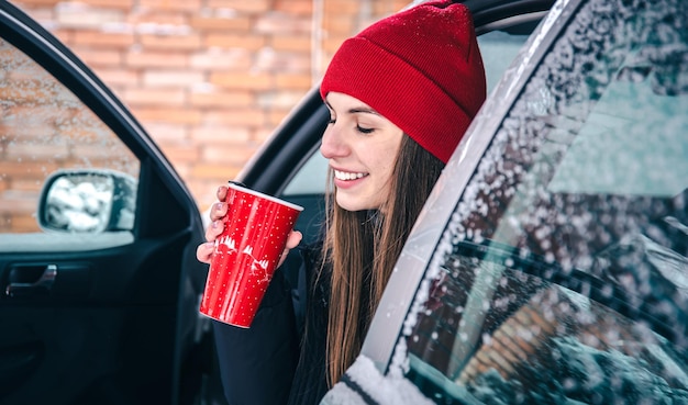 Felice giovane donna con una tazza termica rossa si siede in un'auto in inverno