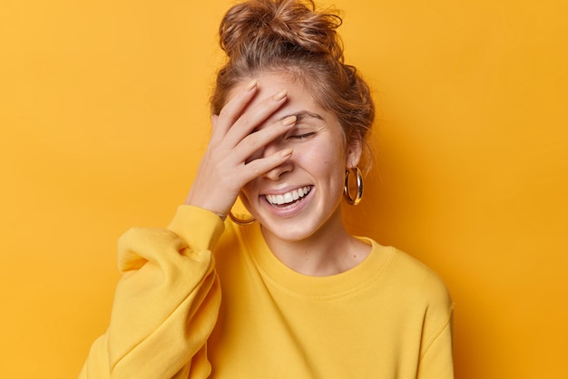 Felice giovane donna copre metà del viso con la mano sorride gioiosamente sorride a denti stretti ed esprime emozioni positive chiude gli occhi dalla gioia isolati su sfondo giallo si erge in posa rilassata