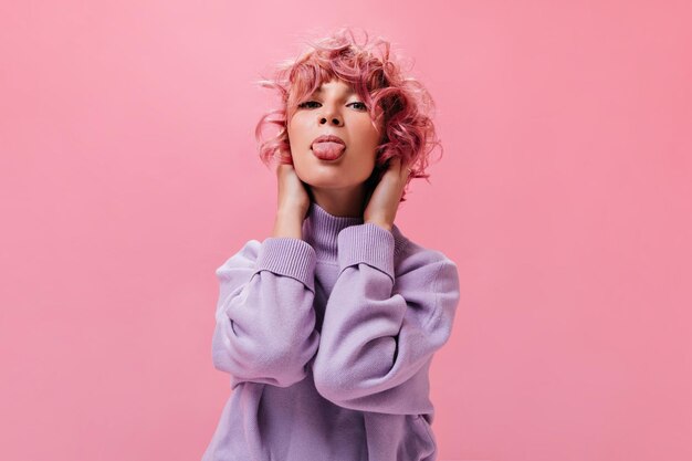 Felice giovane donna dai capelli rosa ricci dimostra la lingua Bella ragazza in maglione viola fa una faccia buffa isolata