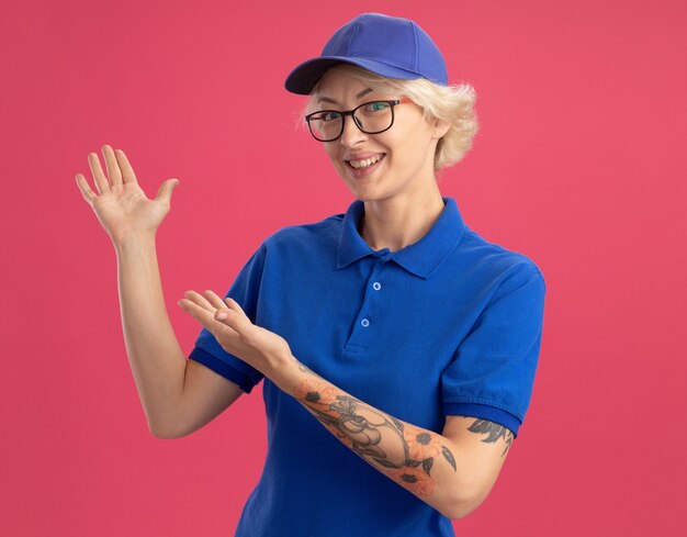 Felice giovane donna delle consegne in uniforme blu e berretto che sorride allegramente presentando lo spazio della copia con le braccia delle sue mani sul muro rosa
