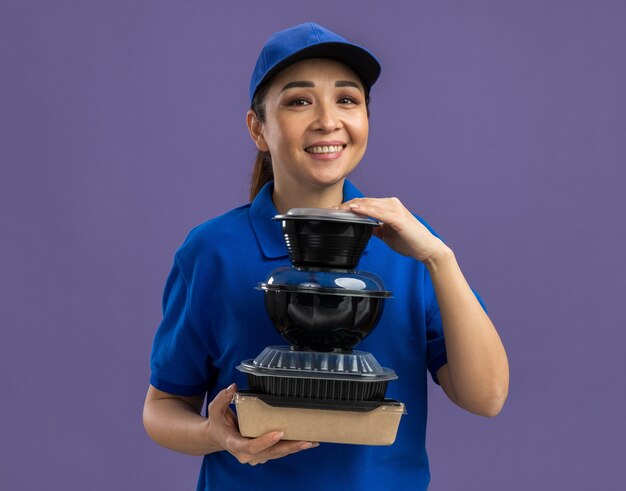 Felice giovane donna delle consegne in uniforme blu e berretto che tiene una pila di confezioni di cibo sorridendo allegramente