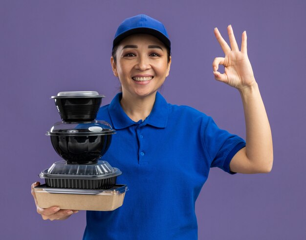 Felice giovane donna delle consegne in uniforme blu e berretto che tiene una pila di pacchi di cibo sorridendo allegramente mostrando segno ok