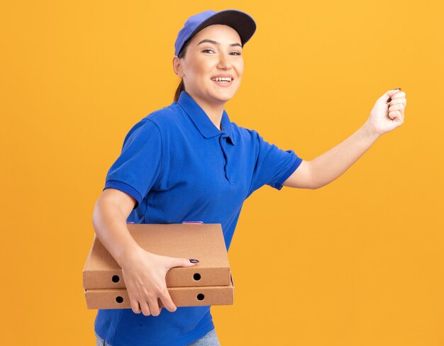 Felice giovane donna delle consegne in uniforme blu e cappuccio in corsa per la consegna di scatole per pizza per il cliente oltre la parete arancione