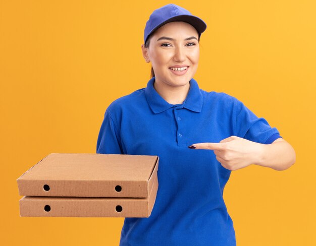 Felice giovane donna delle consegne in uniforme blu e cappuccio tenendo le scatole per pizza puntando con il dito indice verso di loro guardando davanti sorridendo allegramente in piedi sopra la parete arancione