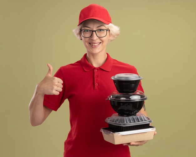 Felice giovane donna delle consegne in uniforme rossa e berretto con gli occhiali che tengono pila di confezioni di cibo sorridente fiducioso che mostra i pollici in su in piedi sopra la parete verde
