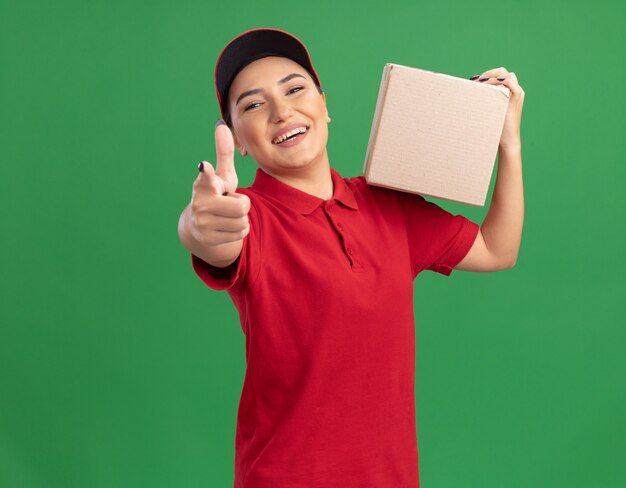 Felice giovane donna delle consegne in uniforme rossa e cappuccio che tiene la scatola di cartone che punta con il dito indice davanti sorridendo allegramente in piedi sopra la parete verde