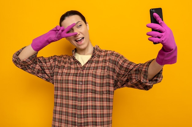 Felice giovane donna delle pulizie in camicia a quadri in guanti di gomma che tiene smartphone facendo selfie sorridente mostrando il segno a V sopra gli occhi in piedi sul muro arancione