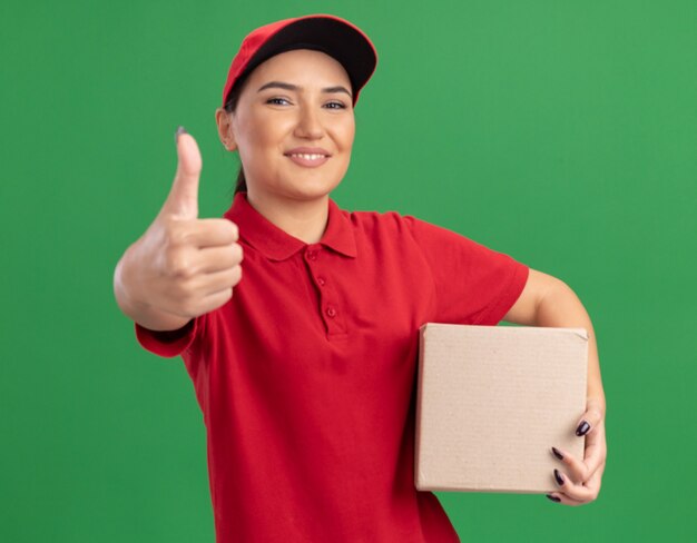 Felice giovane donna di consegna in uniforme rossa e berretto che tiene la scatola di cartone guardando davanti sorridendo allegramente mostrando i pollici in su in piedi sopra la parete verde