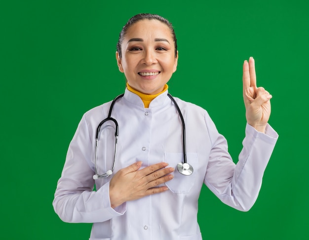 Felice giovane donna dottore in camice bianco con stetoscopio intorno al collo facendo giuramento giuramento giurando con la mano sul petto e mostrando le dita in piedi sul muro verde
