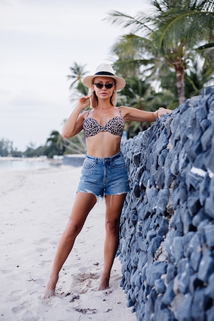 Felice giovane donna europea in pantaloncini di jeans bikini top leopardo e cappello bianco classico sulla spiaggia esotica tropicale sorridente in posa divertendosi Ragazza giocosa sul concetto di viaggio di vacanza