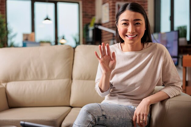 Felice giovane donna filippina cinese asiatica seduta sul divano a casa tenendo il telefono sullo schermo agitando la mano videochiamando amico a distanza online nell'app di chat mobile utilizzando l'applicazione di videochat per smartphone