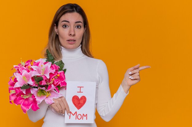 felice giovane donna in dolcevita bianco azienda biglietto di auguri e bouquet di fiori