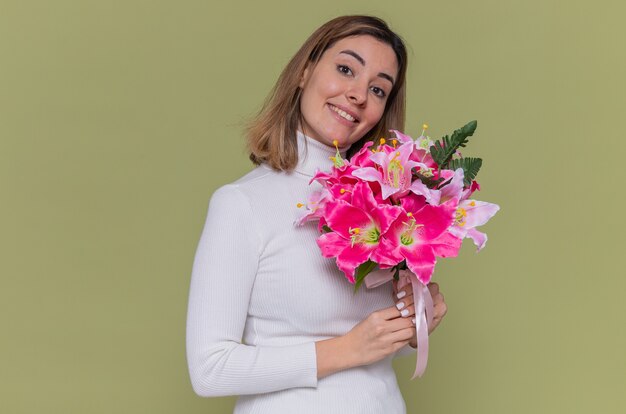 Felice giovane donna in dolcevita bianco azienda bouquet di fiori guardando davanti sorridente allegramente per celebrare la giornata internazionale della donna in piedi sopra il muro verde