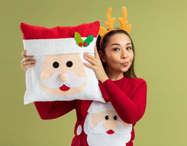 Felice giovane donna in maglione rosso di natale che indossa un bordo divertente con corna di cervo che tengono un cuscino di natale sorridente in piedi sul muro verde