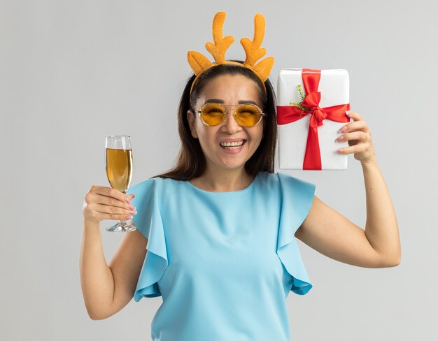 Felice giovane donna in top blu indossando orlo divertente con corna di cervo e bicchieri gialli tenendo un bicchiere di champagne e regalo di Natale sorridendo allegramente