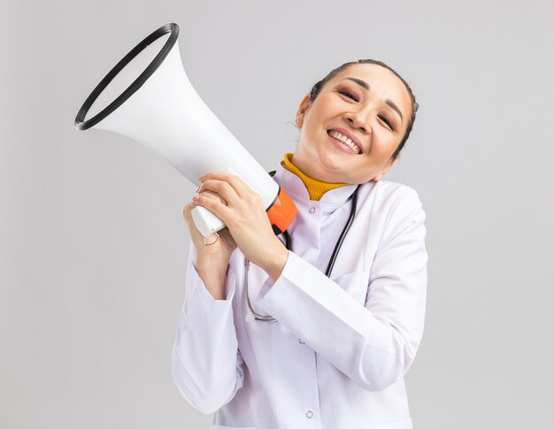 Felice giovane donna medico in camice bianco medico con stetoscopio intorno al collo tenendo il megafono felice e positivo sorridente allegramente in piedi sul muro bianco